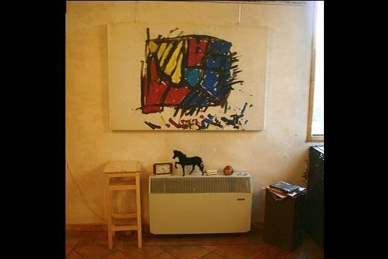 Grande tela chiara appesa sopra un termosifone: macchie di colore rosso, giallo e blu racchiuse da pennellate nere spezzate, con sgocciolature in basso. Sotto, un piccolo cavallo nero di legno e una sveglia.