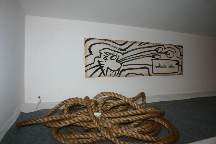 Lungo pannello orizzontale appeso a una parete bianca: su fondo chiaro, grosse linee nere disegnano una figura di profilo con occhi tondi e una bocca aperta; a destra un cartiglio con la scritta a mano «non ti scordar di loro». Sul pavimento grigio, una grossa matassa di corda.