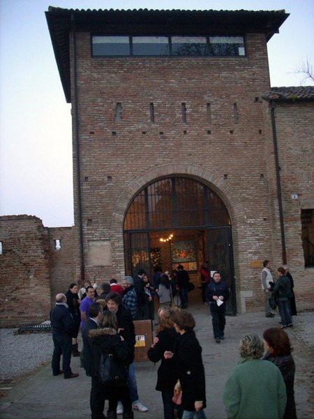 Gente radunata davanti alla torre di mattoni della Porta degli Angeli, sotto l'arco d'ingresso illuminato, all'imbrunire.
