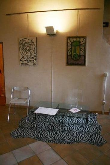 Sala con pareti color crema e pavimento in cotto: due quadri appesi a fili, uno con una figura a linee nere su fondo chiaro e uno verde e rosso; sotto, un tavolino di vetro su un tappeto zebrato e due sedie pieghevoli.