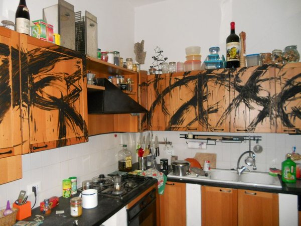 La cucina di casa con i pensili e le ante in legno arancio coperti da grandi pennellate nere curve; sotto, il piano di lavoro con fornelli, lavello e bottiglie.