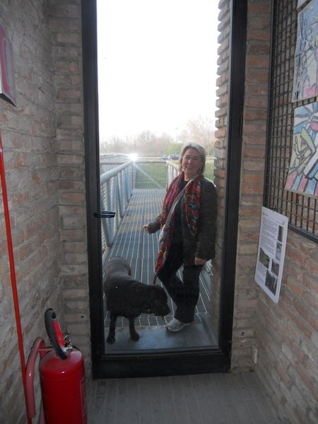 Una donna con sciarpa rossa e un cane nero sulla passerella metallica d'ingresso, tra muri di mattoni; a sinistra un estintore rosso.