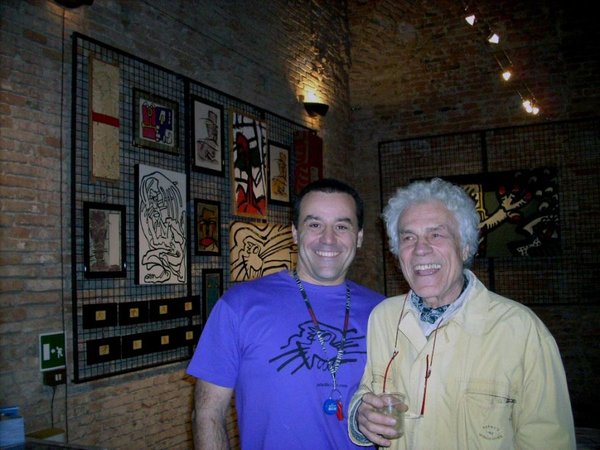 Due uomini sorridenti in posa davanti a una parete di quadri appesi a una griglia: uno in maglia viola con un disegno nero, l'altro anziano, capelli bianchi e giacca chiara, con un bicchiere in mano.