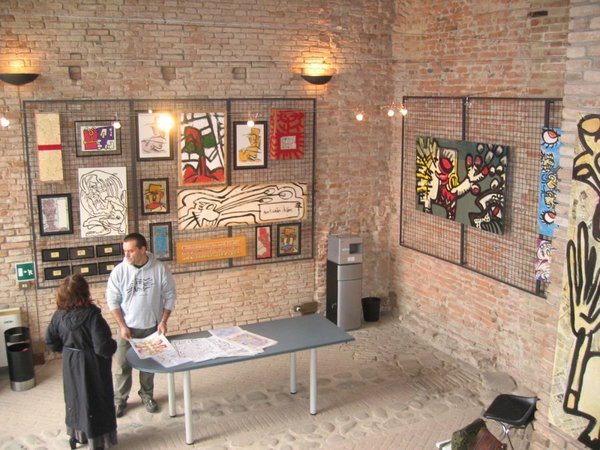 La sala con le pareti di mattoni piene di quadri su griglie; al centro un uomo in felpa grigia e una donna in nero guardano stampe sparse su un tavolo.