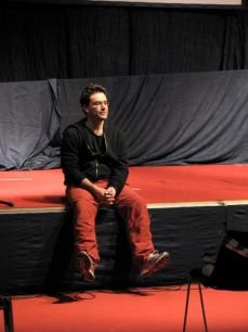 Un uomo con barba corta, maglia nera e pantaloni rossi, seduto sul bordo di un palco rosso davanti a un sipario grigio.