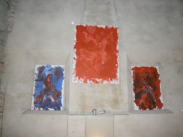 Tre tele su una parete chiara a nicchie: al centro una grande tela rossa, ai lati due più piccole, una blu e rossa e una rosso scuro, dipinte a larghe pennellate.