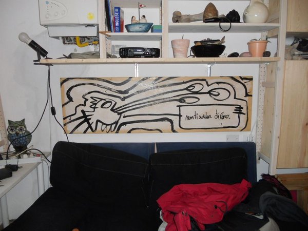 Il pannello «Non ti scordar di loro» in una casa, appeso sopra un divano nero sotto una mensola piena di oggetti: vasi, una lampada, una civetta di legno a sinistra.
