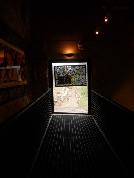 Un corridoio buio con passerella a griglia che porta verso una porta luminosa, sopra la quale è appesa un'opera con pesci neri e gialli.