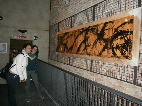 Due ragazze abbracciate sorridono in un passaggio; accanto, appese a una griglia, le ante di cucina dipinte a segni neri su legno arancio, in una lunga cornice chiara.