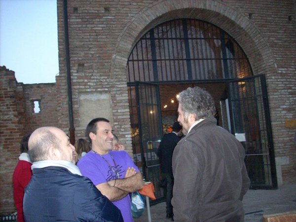 Tre uomini chiacchierano davanti all'arco a cancellata dell'ingresso; quello al centro, in maglia viola, sorride a braccia conserte.