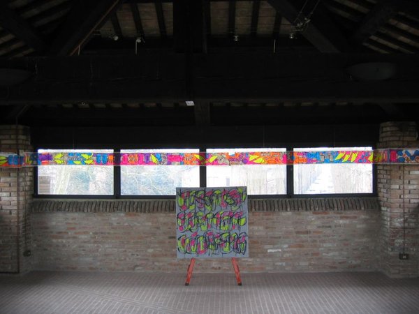 Sala all'ultimo piano con travi scure: una lunga striscia dai colori fluo corre sotto le finestre e al centro, su un cavalletto arancione, una tela con scritte fluo verdi e fucsia.