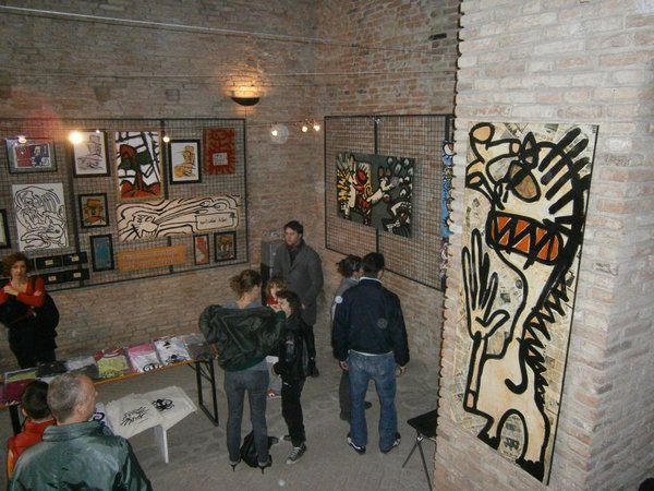 Visitatori nella sala tra pareti di mattoni coperte di quadri; a destra una grande figura nera e bianca con occhiali arancioni e una mano aperta.