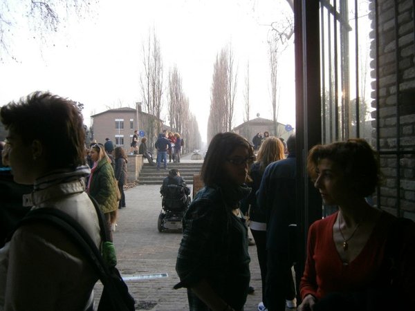 Persone in controluce all'ingresso, sullo sfondo un viale alberato con altre persone e una carrozzina.