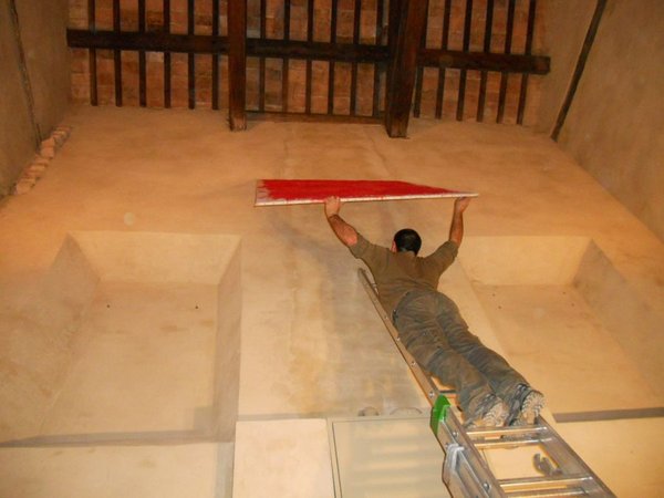 Un uomo sdraiato su una scala appoggiata in orizzontale, sotto il soffitto a travi, sistema una lunga striscia rossa.