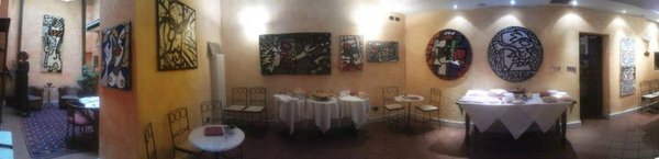 Foto panoramica della sala dell'albergo: pareti color crema con molti quadri, due grandi tondi in bianco e nero, tavoli apparecchiati con tovaglie bianche.