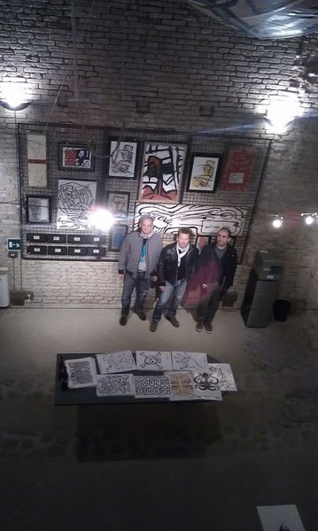 Tre uomini in piedi davanti a una parete di quadri, fotografati dall'alto; in primo piano un tavolo con disegni in bianco e nero.