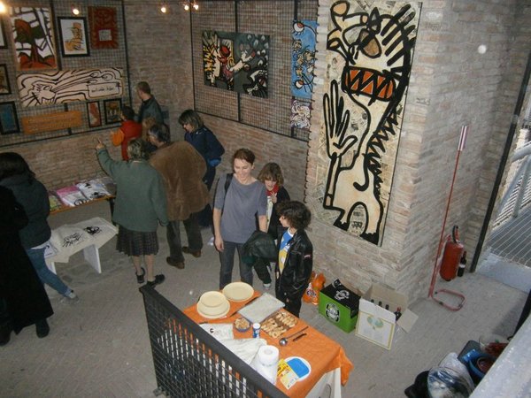 La sala vista dall'alto: pareti di mattoni con quadri su griglie, una grande figura dipinta in bianco, nero e rosso, persone in piedi e un tavolo con dolci in primo piano.