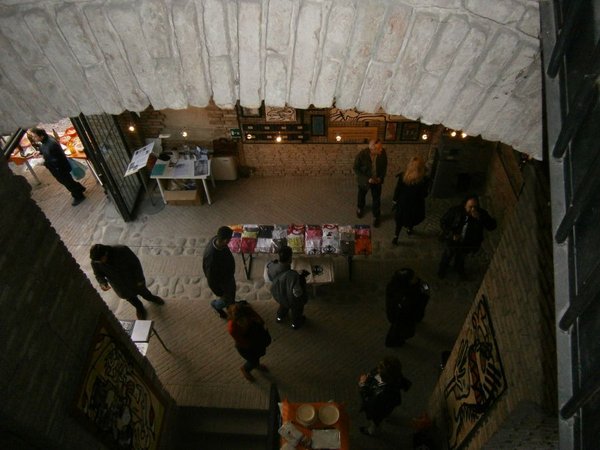 La sala vista dall'alto attraverso un arco: persone che girano tra tavoli con stampe e magliette, quadri alle pareti.