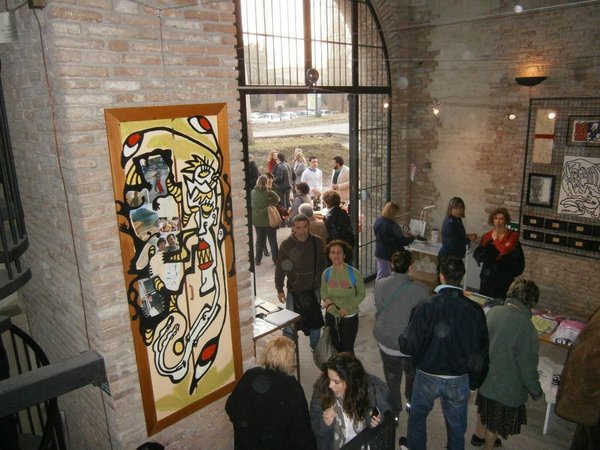 Visitatori nella sala vista dall'alto; a sinistra un'anta d'armadio dipinta con una figura e fotografie incollate, in fondo l'ingresso ad arco aperto sulla luce.