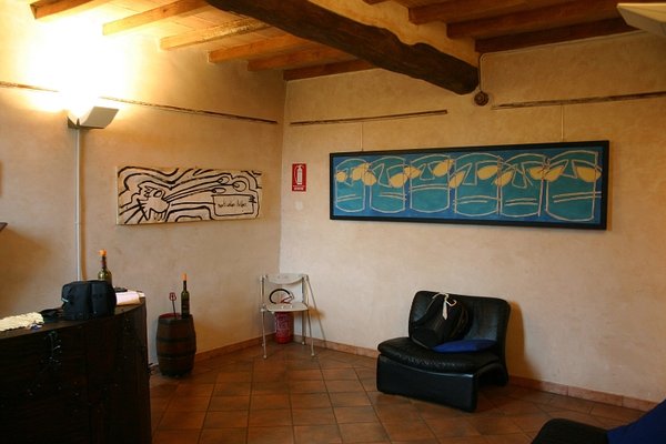 Sala con soffitto a travi di legno e pavimento in cotto: a sinistra un pannello chiaro con figura a linee nere e cartiglio, a destra un lungo quadro blu-verde con una fila di pesci gialli stilizzati; sotto, una poltrona di pelle nera e una sedia.