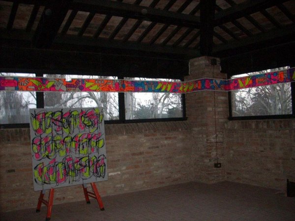 La stessa sala sotto il tetto in penombra: la striscia fluo lungo le finestre e la tela con scritte colorate sul cavalletto.