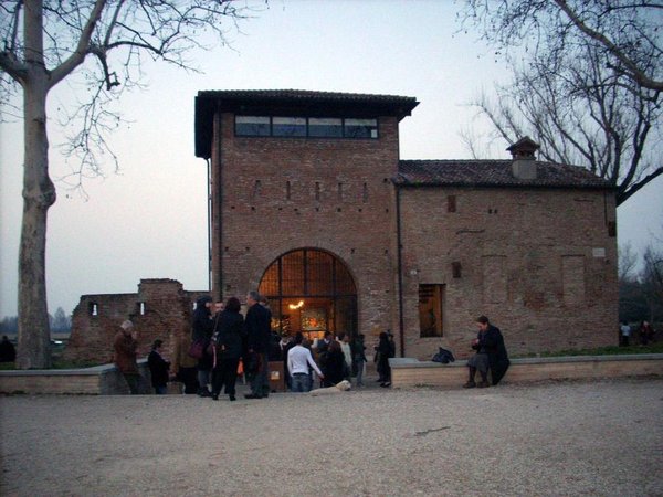 La Porta degli Angeli al tramonto: edificio di mattoni con torre e grande arco illuminato, persone radunate davanti all'ingresso, alberi spogli.
