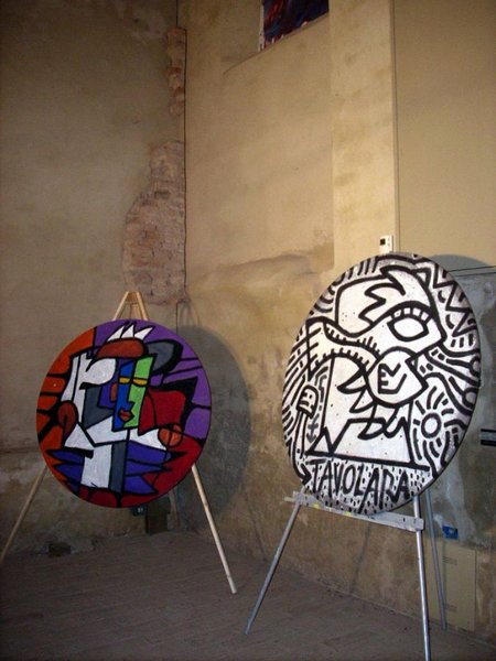 Due tondi dipinti su cavalletti, in una stanza dai muri antichi: uno a campiture rosso, blu e bianche, l'altro in bianco e nero con volti e la scritta «STAVOLA ART».