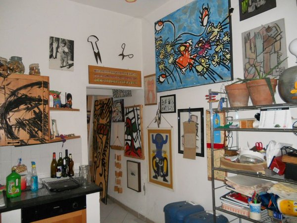 Un angolo della cucina con le pareti piene di quadri: in alto un grande quadro azzurro con pesci colorati, un volto in cornice, figure su tavole; a sinistra un'anta dipinta a segni neri, a destra una scaffalatura metallica.